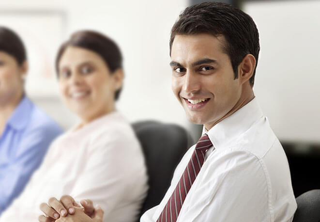 Smiling professional man in a white shirt and tie, sitting in a meeting with two colleagues in the background.